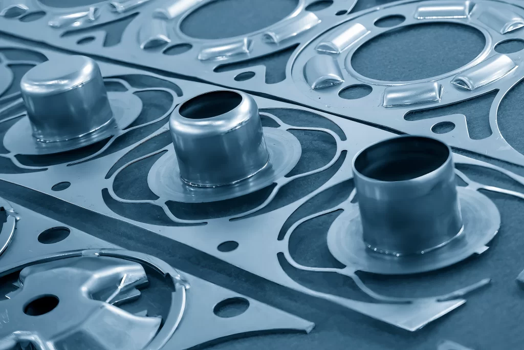 Close-up de peças metálicas técnicas com repuxo profundo, recém-fabricadas e ainda presas ao esqueleto da chapa após o processo de estampagem de precisão.